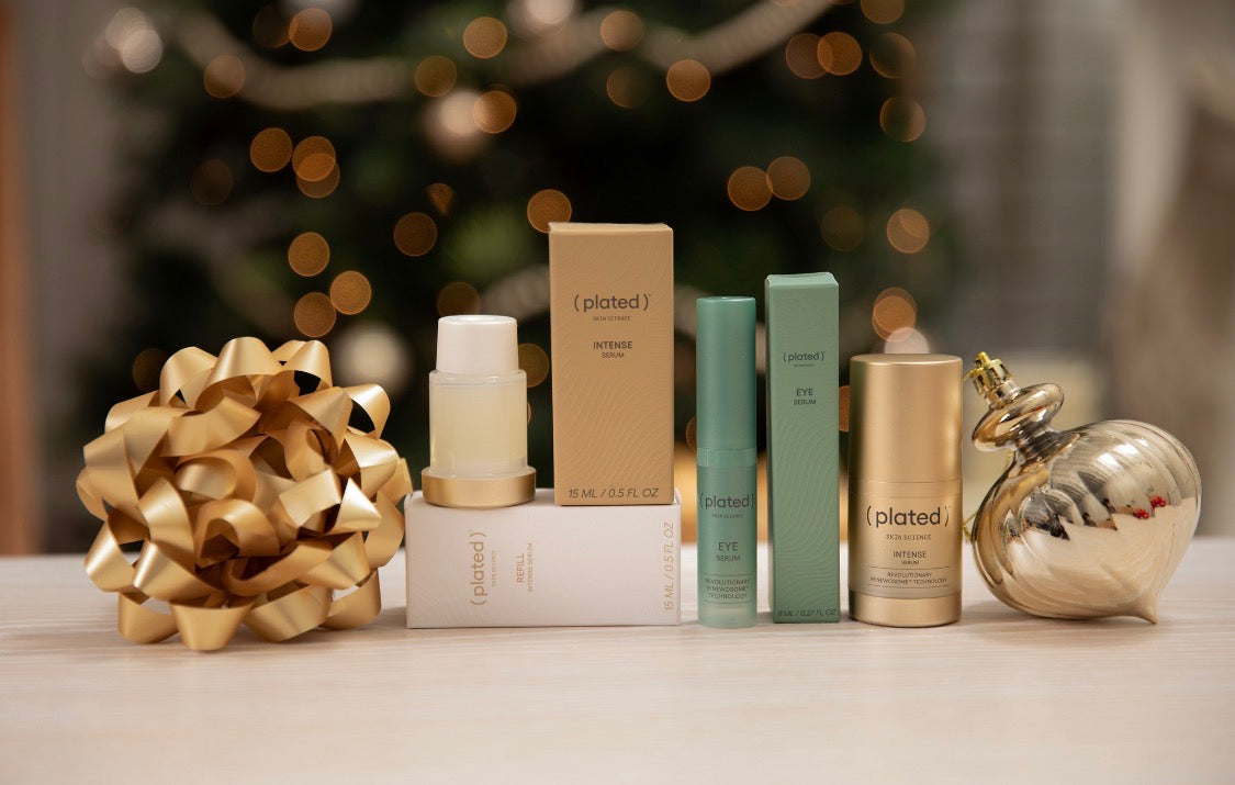 ( plated )™ INTENSE Serum, EYE Serum, and Refill displayed with a gold holiday bow and ornament in front of a softly lit Christmas tree.